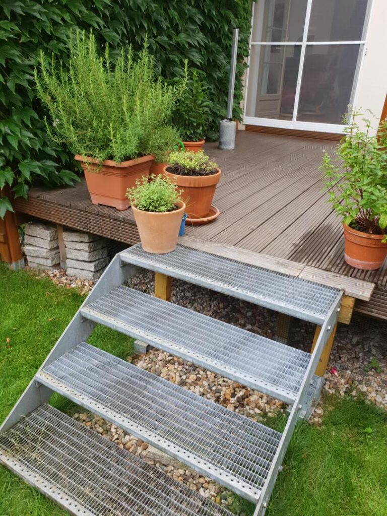 Metalltreppe führt zu Holzterrasse mit Kräutertöpfen.